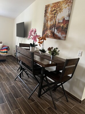 A dining area with a wooden table and chairs, a flat-screen TV mounted on the wall, and a painting depicting a landscape.