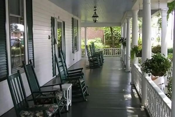 A covered porch with white columns and green shutters, featuring a row of chairs and a table.