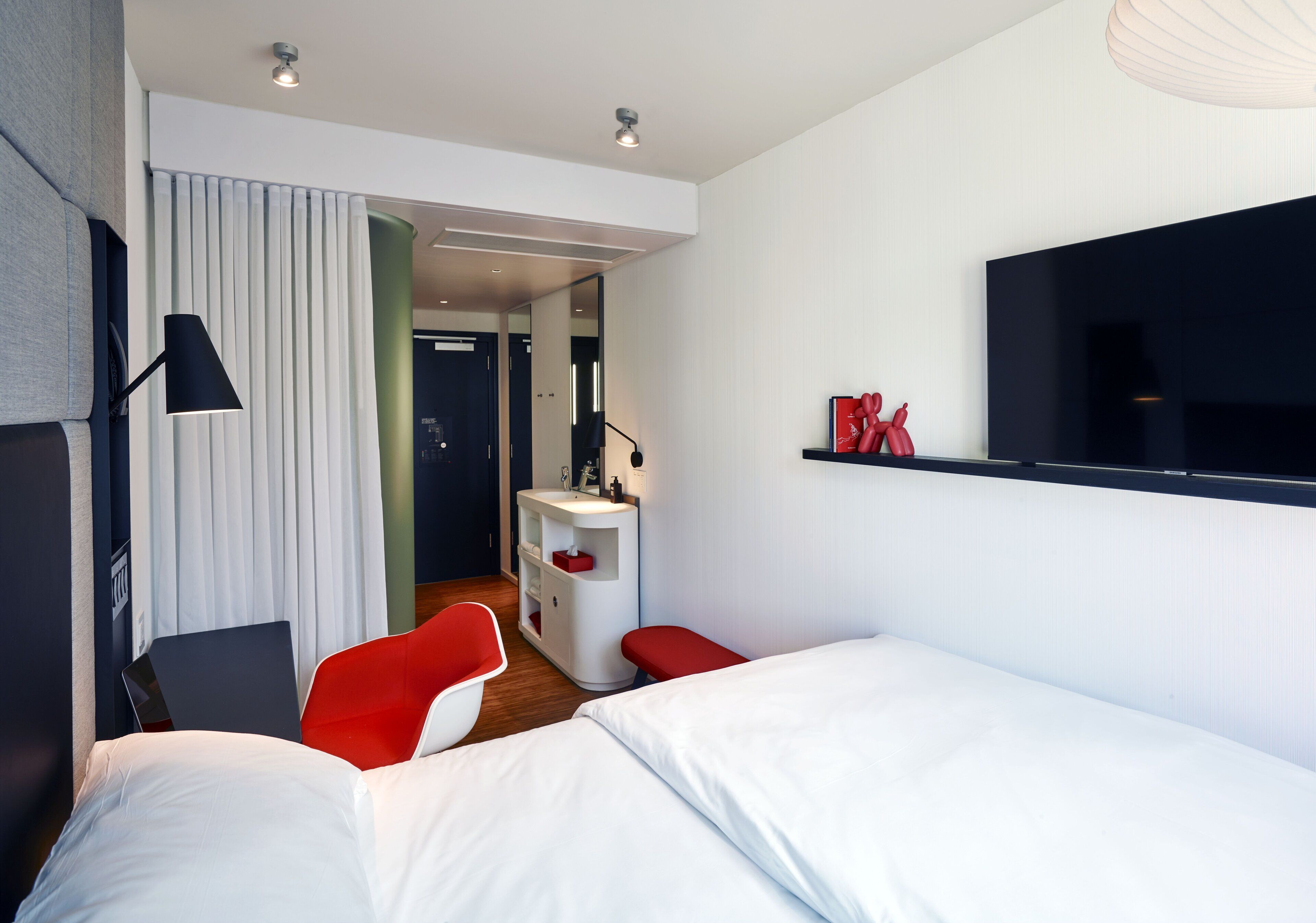 Moderni hotellihuone, jossa on sänky, työpöytä tuolilla, seinälle asennettu televisio ja kylpyhuone, joka näkyy oven läpi.