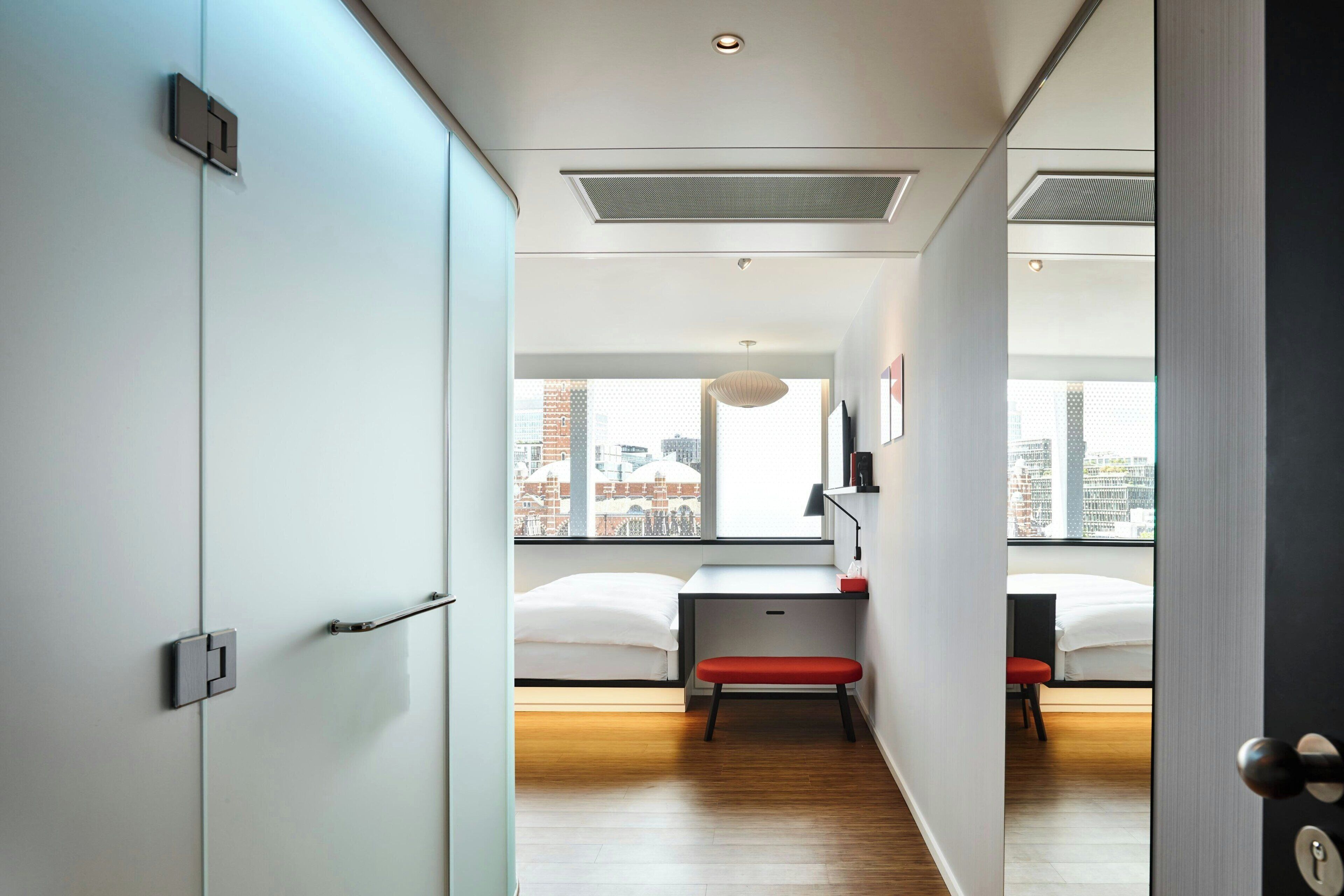 A modern hotel room with a large window, a desk, a red bench, and a bed.