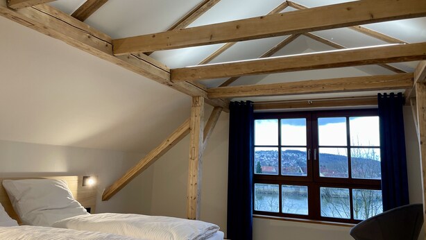 Ein Schlafzimmer mit schrägen Wänden, einem großen Fenster mit Vorhängen, einem Bett mit weißer Bettwäsche und einem Sessel.