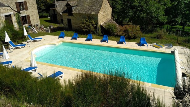Una piscina rectangular con sillas reclinables azules y sombrillas, rodeada de edificios de piedra y vegetación.