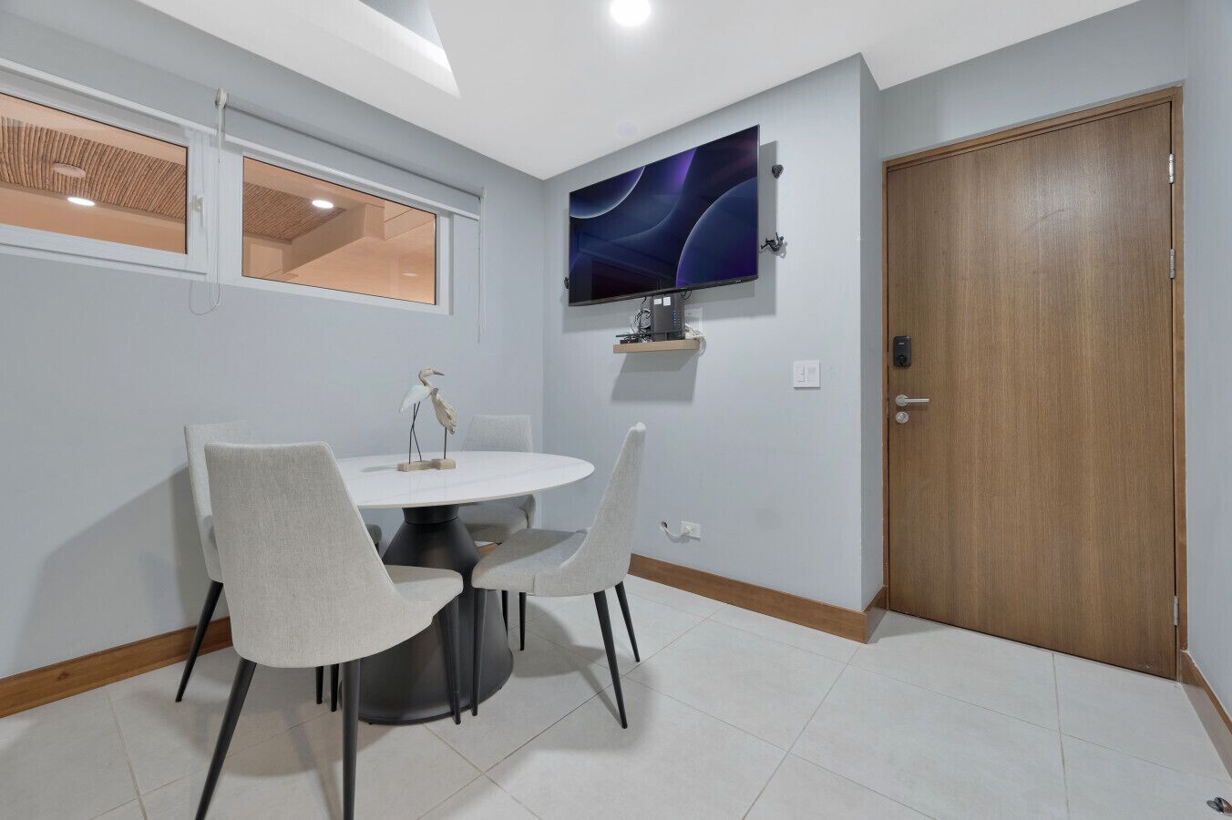 A dining area with a round table, four chairs, a TV mounted on the wall, and a wooden door.