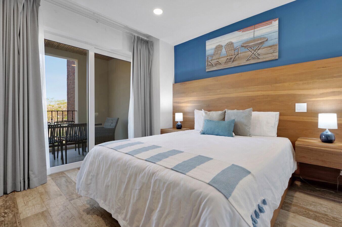 Une chambre d’hôtel avec un grand lit, une tête de lit en bois, un balcon avec des chaises et un tableau au mur.