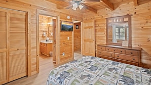 A wooden cabin bedroom with a bed, a ceiling fan, a dresser, and a bathroom with a sink and mirror. - SMOKY MTN STAYS OFF LAKE w/POOL & HOT TUB (Sevierville)