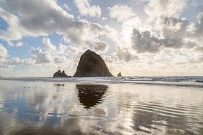 Una playa con aguas tranquilas que reflejan el cielo y las nubes, y una formación rocosa destacada en la distancia.