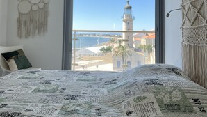 A bedroom with a large window, a bed with a patterned bedspread, a macrame wall hanging, and a view of a lighthouse and beach.