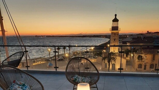 A terrace with a view of the sea and a lighthouse, featuring wicker chairs, a table with a lamp, and a patterned rug.