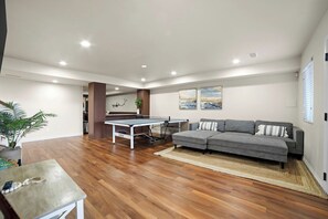 A spacious living area with a ping pong table, grey sofa, and wooden flooring.