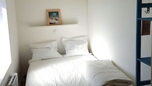 Una habitación pequeña con cama individual, cubrecama blanco, un cuadro enmarcado en la pared y una estantería azul a la derecha.