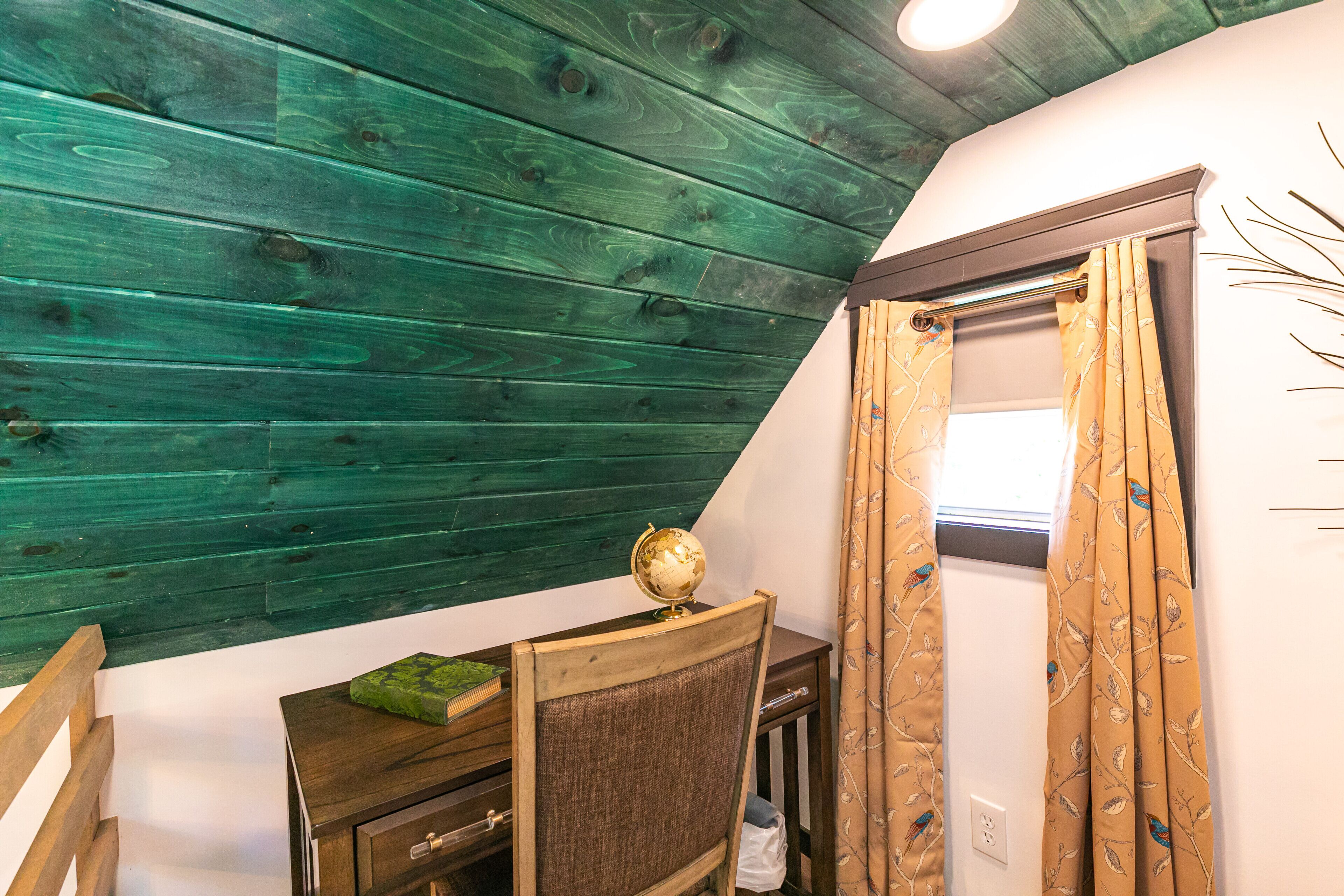 A room with a sloped ceiling, a wooden desk with a globe, a chair, and a window with patterned curtains.