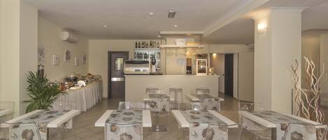 A restaurant with clear glass tables and chairs, a bar area with bottles, and a kitchen in the background.