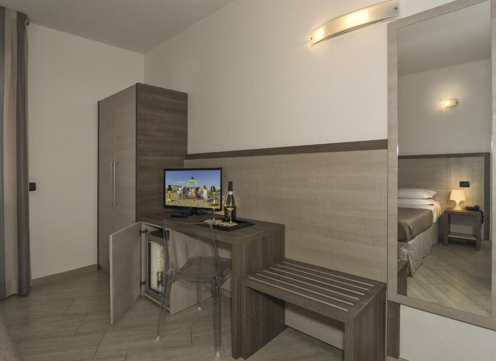 A hotel room with a desk, a chair, a small refrigerator, a bed, a bedside table with a lamp, and a large mirror.