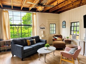Una sala de estar acogedora con un sofá gris oscuro, una mesita de centro redonda, un sillón rosa y un sillón marrón. Un cartel con la palabra 'Welcome' está en la pared, y hay ventanas grandes que dan a vistas de zonas verdes.