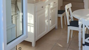 Een eetruimte met een witte tafel en stoelen, een wit kastje met lades, en een trap naar een bovenverdieping.