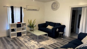En stue med en mørkeblå sofa, et glas sofabord, en potteplante og en hvid aircondition-enhed på væggen.