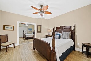 Une chambre à coucher avec un lit à baldaquin en bois, un ventilateur de plafond et une vue sur la plage, visible à travers un miroir.