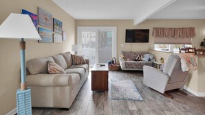 Ein Wohnzimmer mit einem beigen Sofa, einem Holztisch, einem gemusterten Teppich und einem an der Wand befestigten Fernseher.