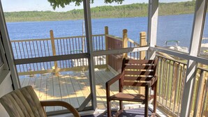 A covered outdoor seating area with a view of a lake, wooden deck, and a glass railing.
