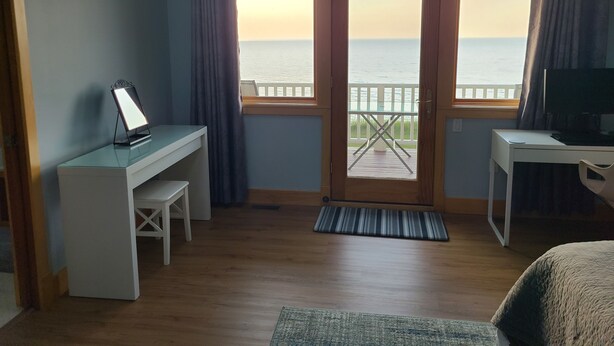 Ein Zimmer mit Schreibtisch, Stuhl, Bett, Fernseher und Meerblick durch eine Tür.