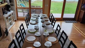 Ein Essbereich mit einem langen Tisch, der für eine Mahlzeit gedeckt ist, großen Fenstern mit Blick ins Freie und einer Küchenzeile inklusive Kaffeemaschine.