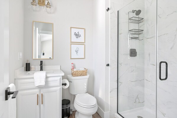 A modern bathroom with a white vanity, a toilet, a shower with a glass door, and framed artwork on the wall. - Brand New Luxury Home less than a mile from Notre Dame (South Bend)