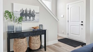 Un couloir avec un escalier, une porte, un tableau encadré, une console ornée d’objets décoratifs et deux paniers en osier.