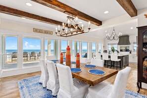 A spacious dining area with a view of the ocean, featuring a wooden table, white chairs, and a chandelier.
