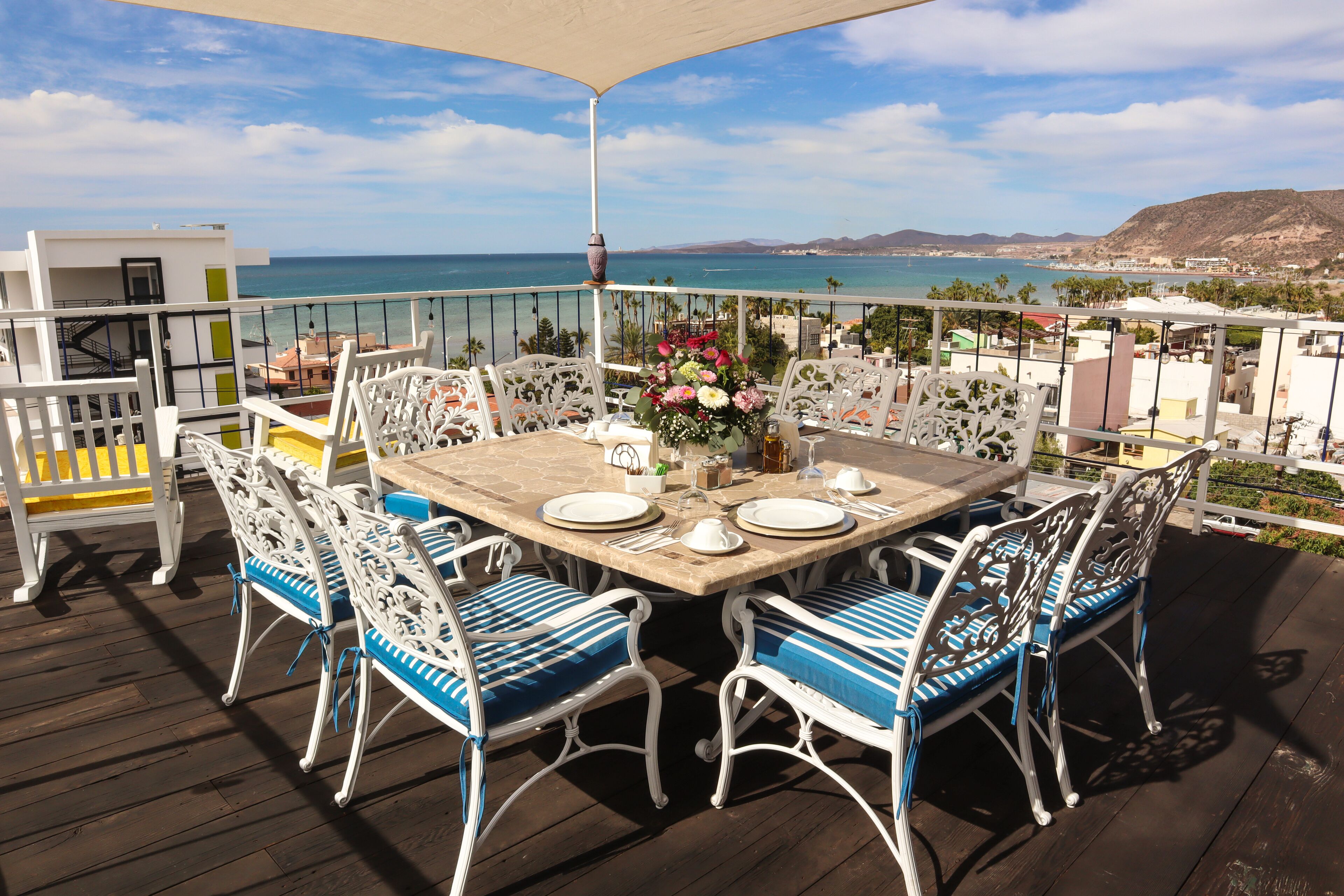 Eine Terrasse mit Tisch, gedeckt für vier Personen, umgeben von weißen Stühlen mit blauen Kissen, mit Blick auf die Küste.