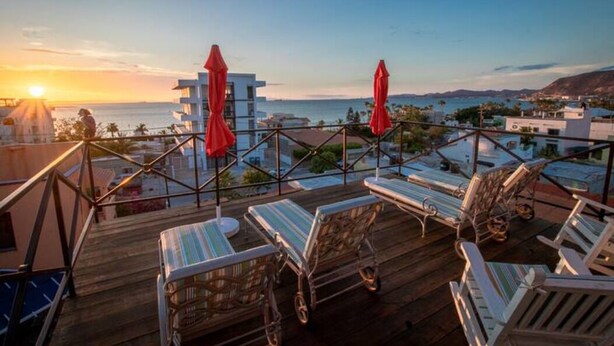 A rooftop terrace with lounge chairs and red umbrellas, overlooking a coastal cityscape during sunset. - Suite Giuseppe, luxury and comfort close to the malecon. Breakfast included  (La Paz)