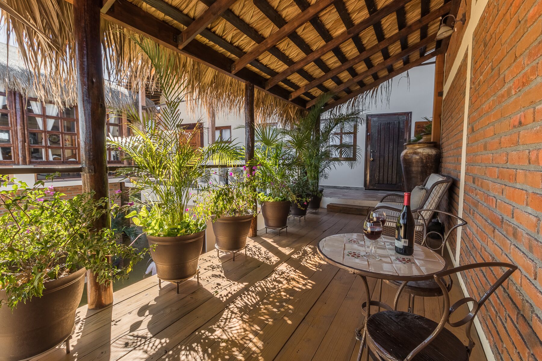 A covered outdoor patio with wooden flooring, potted plants, a round table with chairs, and a bottle of wine.