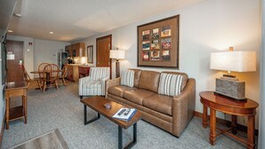A living room with a brown leather sofa, two striped armchairs, a wooden coffee table, and a side table with a lamp. A framed collage hangs on the wall above the sofa. - Suites at Silver Tree: A117 One Bedroom Lake View (Oakland)