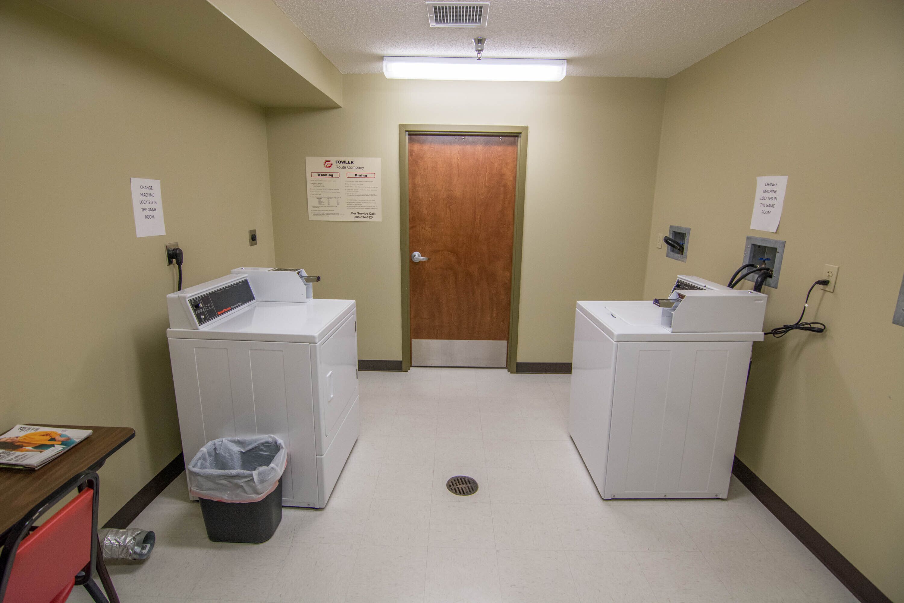 A laundry room with two washing machines, a trash bin, and a door with a sign above it.