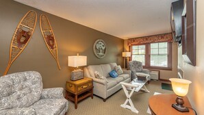 A living room with a sofa, armchair, and a small table with a lamp. There is a wall decoration and a window with blinds. - Suites at Silver Tree: A208 One Bedroom Standard View (Oakland)