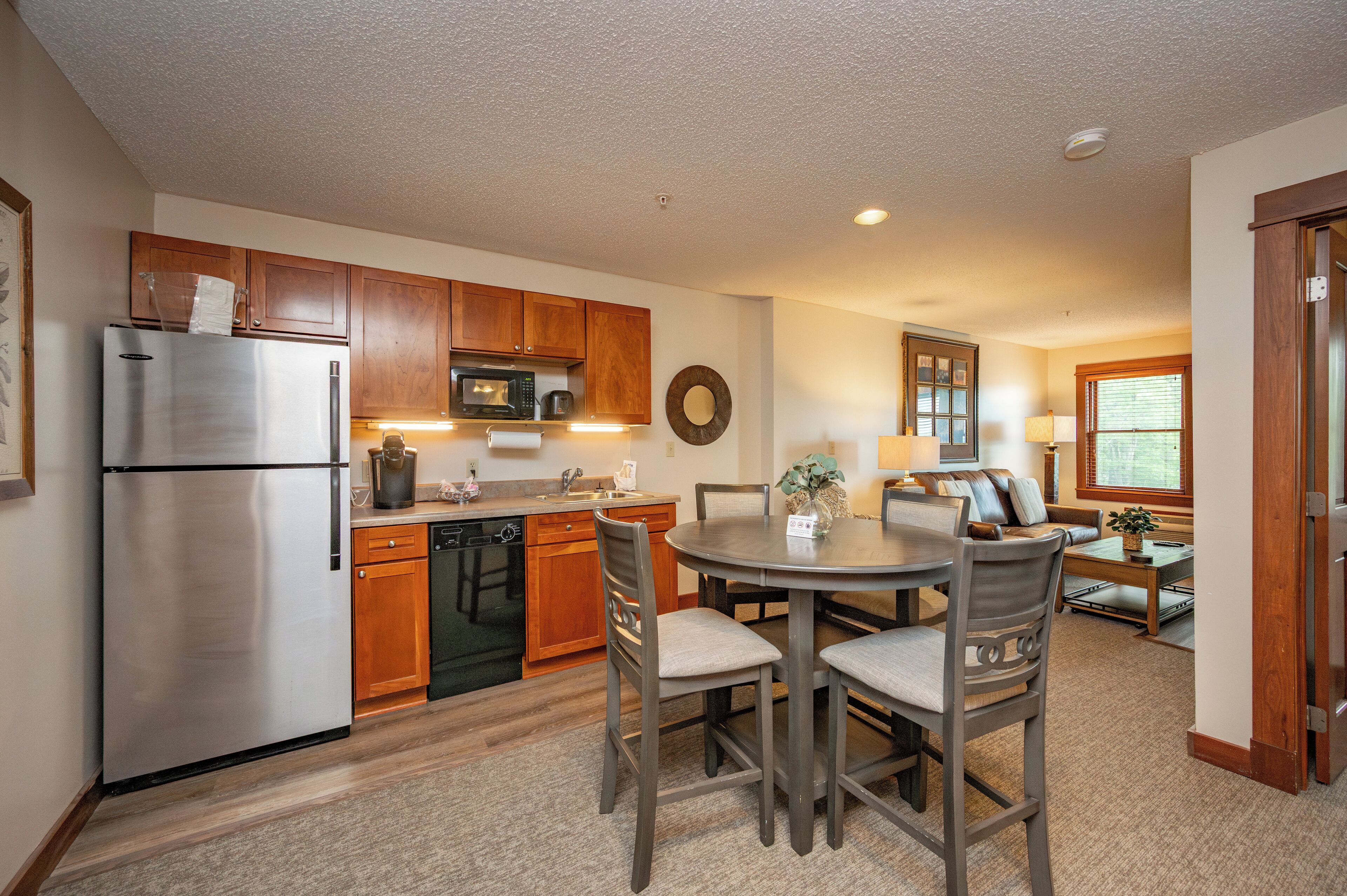 A compact kitchen with wooden cabinets, a stainless steel refrigerator, and a microwave. There is a dining area with a round table and chairs.