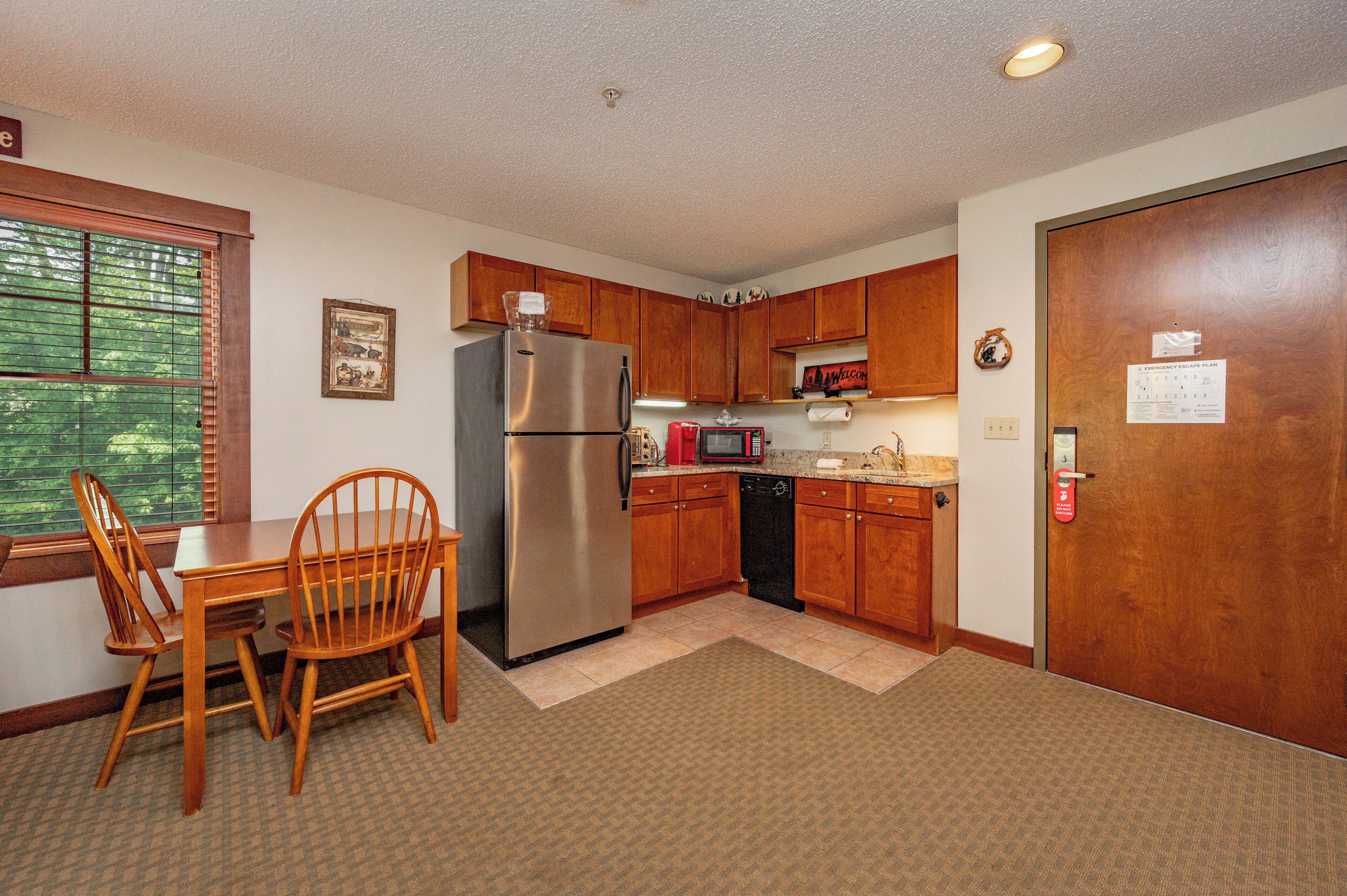 A compact kitchen with a refrigerator, microwave, and dishwasher. There is a dining table with chairs and a window with blinds.