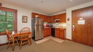 A compact kitchen with a refrigerator, microwave, and dishwasher. There is a dining table with chairs and a window with blinds.
