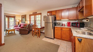 A spacious kitchen with wooden cabinets, a stainless steel refrigerator, a microwave, and a sink. There is a dining area with a table and chairs, and a living room with a sofa and a window. - Suites at Silver Tree: A218 One Bedroom Standard View (Oakland)