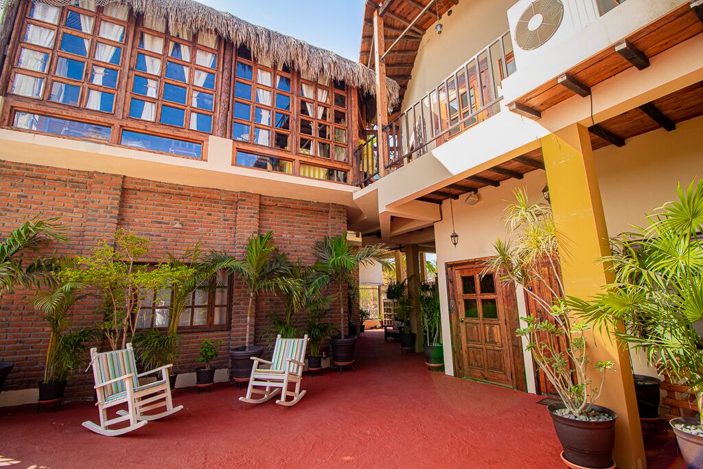 A courtyard with a brick wall, wooden rocking chairs, potted plants, and a thatched-roof building.