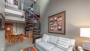 A living room with a grey sofa, wooden dining table, and chairs, a spiral staircase, and a framed collage on the wall. - Suites at Silver Tree: A311 One Bedroom & Loft Lake View (Oakland)