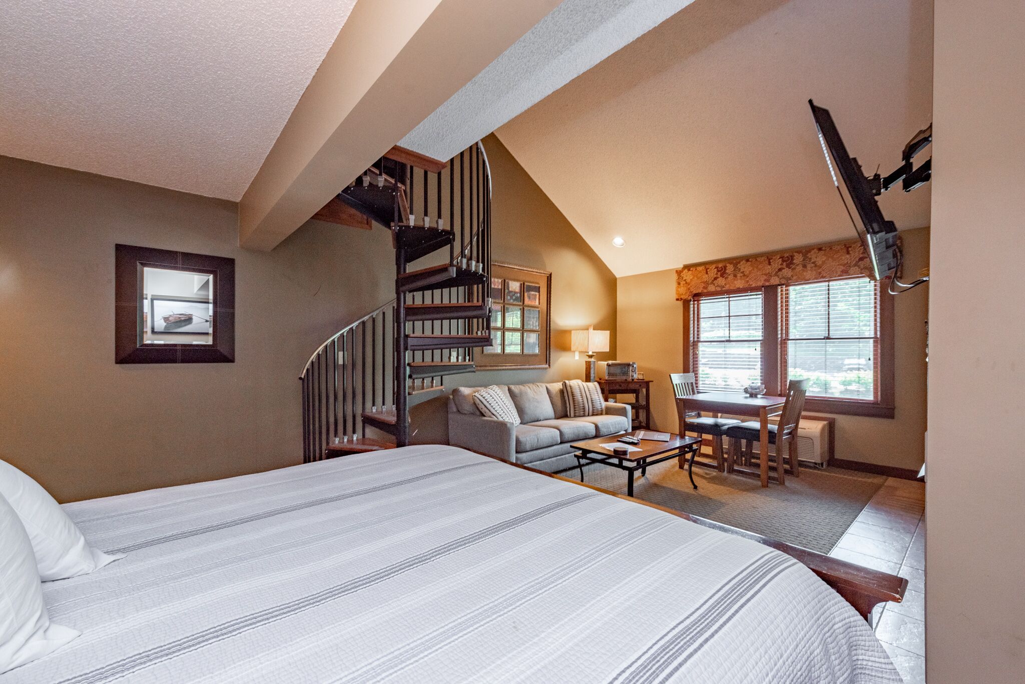 A bedroom with a large bed, a sofa, a dining table, and a staircase leading to an upper level.