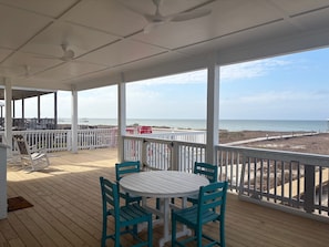 Um espaço ao ar livre coberto, com mesa e cadeiras, vista para a praia e um guarda-corpo branco.