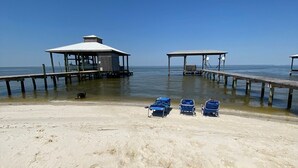 A beach with a wooden pier leading to a covered structure. There are blue beach chairs and a stroller on the sand. - 4BR/3BA with Private Beach, Pier/Boat Lift, Dog Friendly, Free Kayaks, Best WiFi (Dauphin Island)