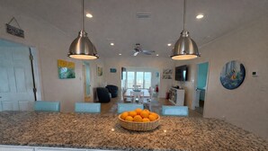 A kitchen with granite countertops, a central island with a basket of oranges, pendant lighting, and a dining area in the background. - 4BR/3BA with Private Beach, Pier/Boat Lift, Dog Friendly, Free Kayaks, Best WiFi (Dauphin Island)