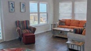 A spacious living room with a large window, a ceiling fan, a colorful striped armchair, a red ottoman, a sofa with orange cushions, and a round rug. - 4BR/3BA with Private Beach, Pier/Boat Lift, Dog Friendly, Free Kayaks, Best WiFi (Dauphin Island)
