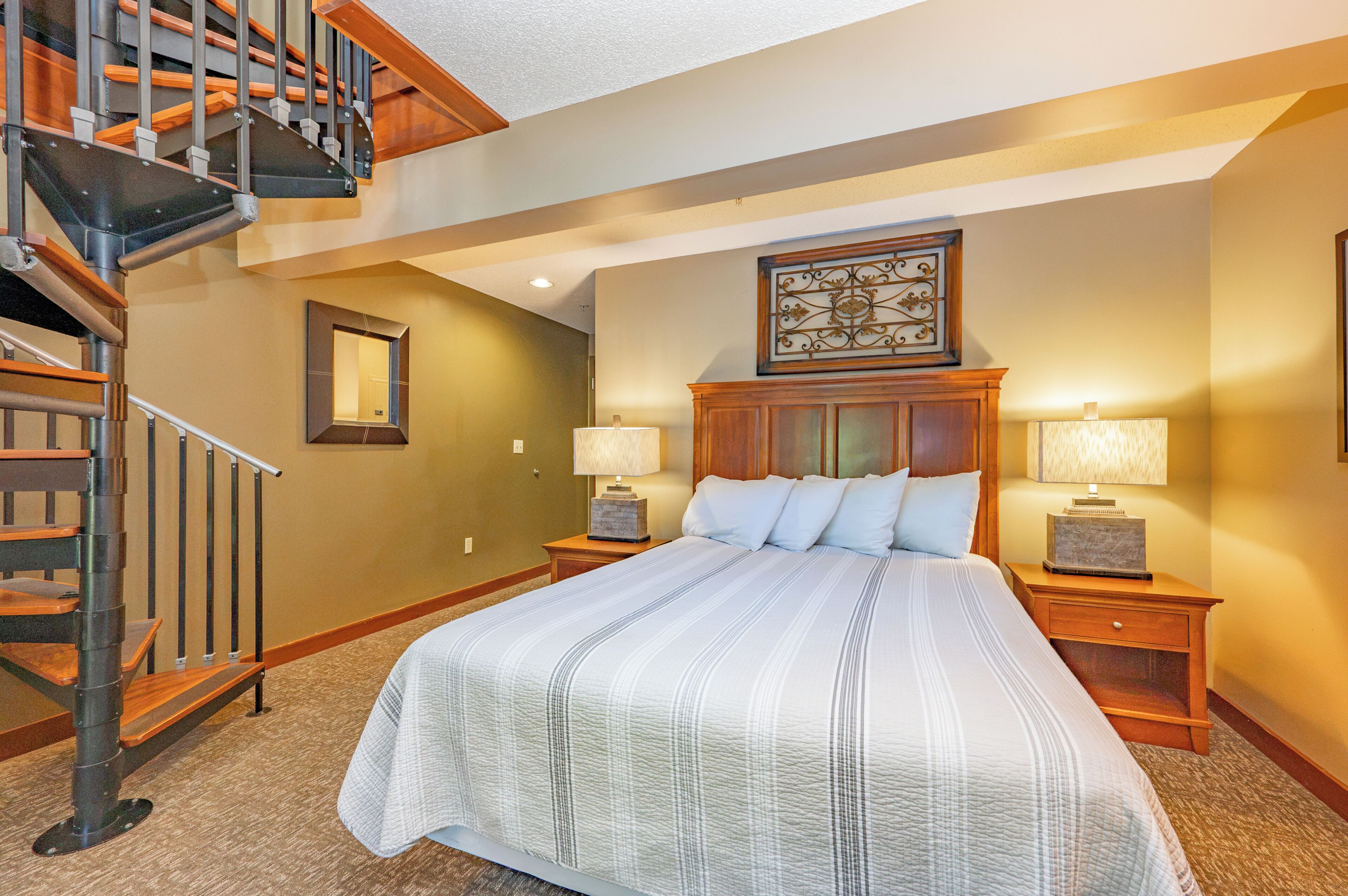 A bedroom with a large bed, two bedside tables with lamps, a wooden headboard, a decorative wall piece, and a staircase.