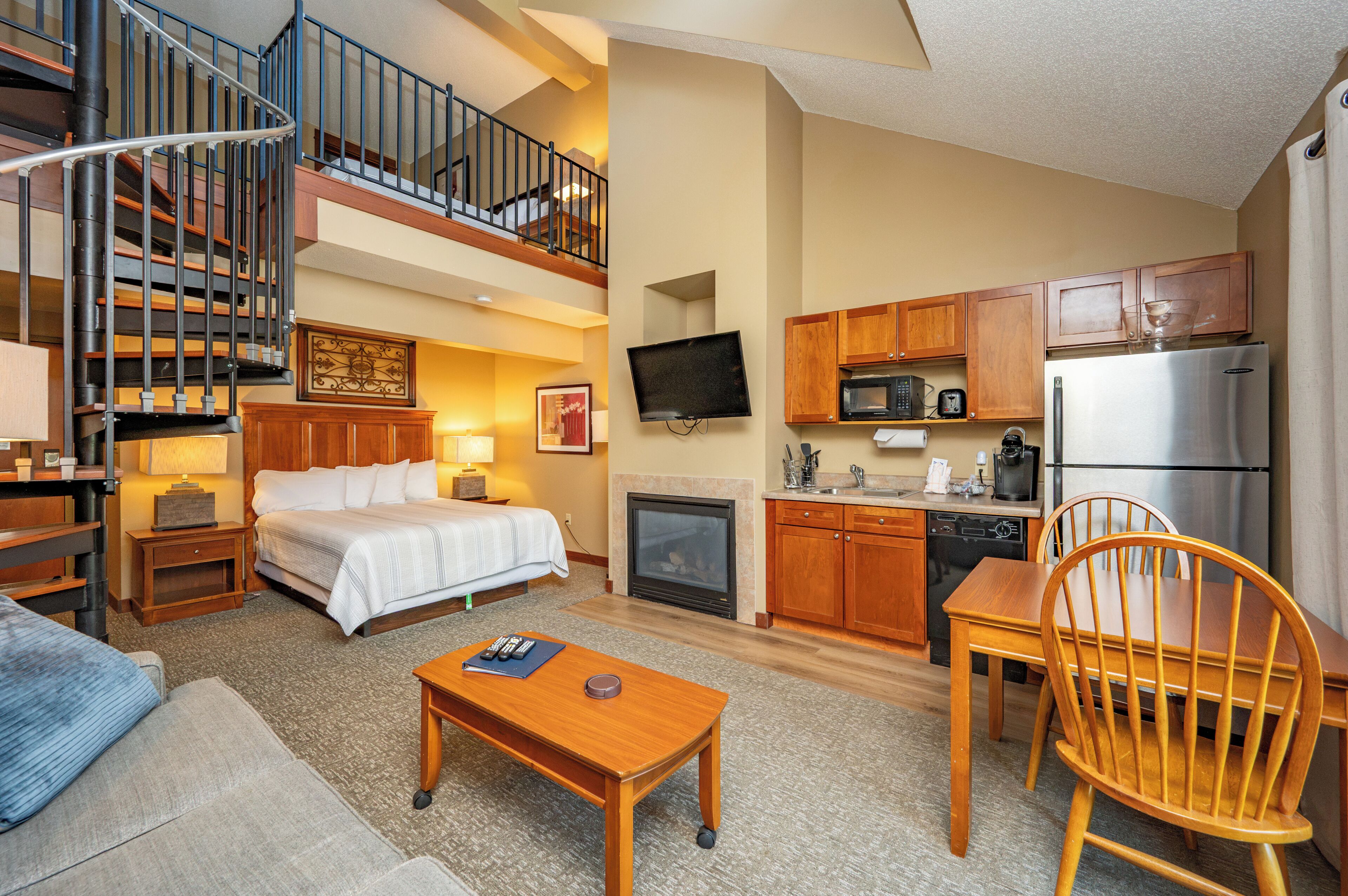 A hotel room with a bed, a desk, a chair, a television, a kitchen area with a microwave, a refrigerator, and a staircase.