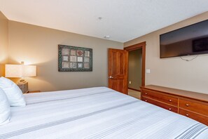 A bedroom with a bed, a nightstand, a dresser, a television, and a door leading to another room. - Suites at Silver Tree: B106 One Bedroom Standard View (Oakland)