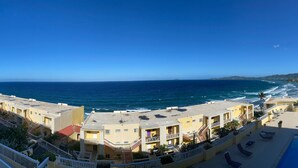 Ein Küstenhotel mit mehreren Balkonen mit Meerblick, einem Swimmingpool und Blick auf den Strand und die entfernten Hügel.