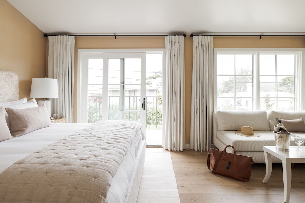 A hotel room with a large bed, a sofa, a small table with a hat and a bottle, and a handbag on the floor.
